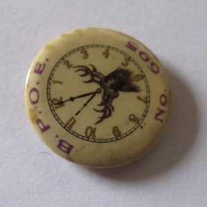 May include: A round, off-white button with a clock design. The clock face has purple numerals and hands, with the text "B.P.O.E." and "SON" in purple. A dark graphic is in the center. The button is slightly worn.