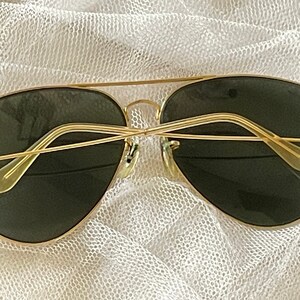 May include: Gold-framed aviator sunglasses with dark green lenses. The sunglasses have a classic double bridge and a sleek, minimalist design. The temples are thin and curve gently for a comfortable fit. A timeless accessory for eye protection.