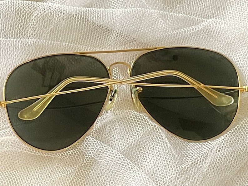 May include: Gold-framed aviator sunglasses with dark green lenses. The sunglasses have a classic double bridge and a sleek, minimalist design. The temples are thin and curve gently for a comfortable fit. A timeless accessory for eye protection.