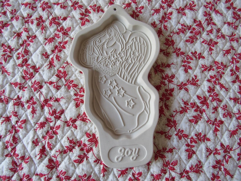 Puede incluir: Un cortador de galletas de cer&aacute;mica blanca con forma de &aacute;ngel sosteniendo una pancarta con estrellas. El cortador tiene la palabra "joy" inscrita en la parte inferior.