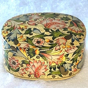 May include: A small, oval-shaped decorative box with a cream-colored base. The box is adorned with a vibrant floral pattern in shades of pink, green, blue, and yellow. The lid and sides are fully covered with the intricate design.