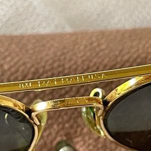 May include: Close-up of gold-rimmed sunglasses with dark lenses. The text "9121 PAT. USA" is visible on the frame. The sunglasses have a classic design and are set against a neutral background.