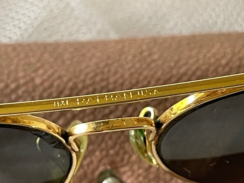 May include: Close-up of gold-rimmed sunglasses with dark lenses. The text "9121 PAT. USA" is visible on the frame. The sunglasses have a classic design and are set against a neutral background.