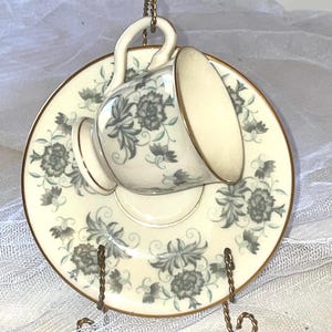 May include: A delicate porcelain teacup and saucer set with a floral design in shades of gray and green. The set is displayed on a gold metal stand.