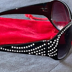 May include: Black sunglasses with dark lenses and a decorative pattern of white rhinestones along the temples. The sunglasses are resting on a red velvet pouch, suggesting a protective case.
