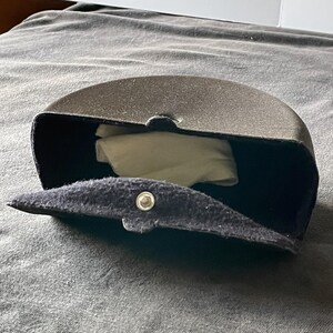 Op de afbeelding: Een donkergrijze, halfronde brillenkoker met een druksluiting. De koker is open en onthult een witte doek binnenin. De koker is gemaakt van een textuurrijk materiaal.