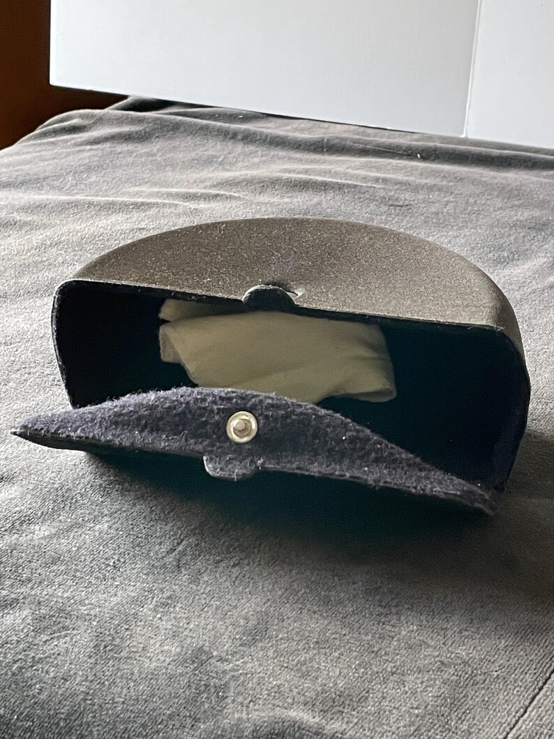Op de afbeelding: Een donkergrijze, halfronde brillenkoker met een druksluiting. De koker is open en onthult een witte doek binnenin. De koker is gemaakt van een textuurrijk materiaal.