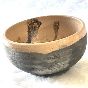 May include: A handmade ceramic bowl with a unique design. The bowl features a two-tone color scheme, with a light tan interior and a dark gray exterior. The rim has a light tan band. The interior has dark brown markings.