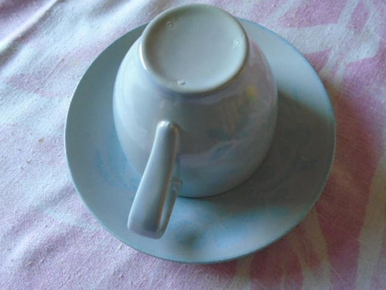 May include: A light blue teacup and saucer set. The cup has a smooth, rounded shape and a small handle. The saucer has a scalloped edge.