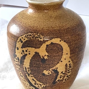 May include: A small, brown ceramic vase with a white rim and a brown and yellow abstract design.