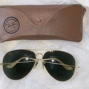 May include: Gold-framed aviator sunglasses with dark green lenses and a brown leather case. The case has a snap closure and a Ray-Ban logo. The sunglasses are a classic style, suitable for various occasions.