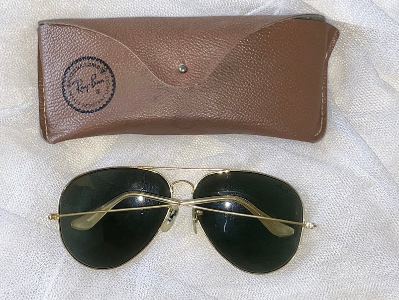 May include: Gold-framed aviator sunglasses with dark green lenses and a brown leather case. The case has a snap closure and a Ray-Ban logo. The sunglasses are a classic style, suitable for various occasions.