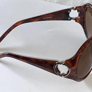 May include: Tortoiseshell sunglasses with brown lenses. The frames have a dark brown pattern and feature silver-colored accents at the temples. The text "JN1210568" is printed on the arm.