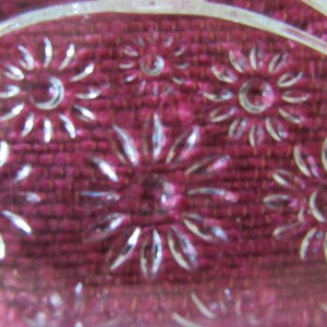 K&ouml;nnte beinhalten: Nahaufnahme einer durchsichtigen Glasschale mit einem dekorativen Muster aus gepr&auml;gten Blumen. Die Schale liegt auf einer burgunderfarbenen Stoffoberfl&auml;che.