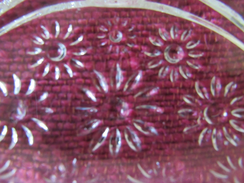 K&ouml;nnte beinhalten: Nahaufnahme einer durchsichtigen Glasschale mit einem dekorativen Muster aus gepr&auml;gten Blumen. Die Schale liegt auf einer burgunderfarbenen Stoffoberfl&auml;che.