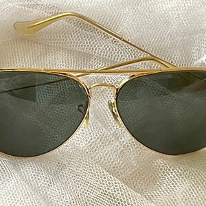 May include: Gold-framed aviator sunglasses with dark green lenses. The sunglasses have a classic teardrop shape and a double bridge. The Ray-Ban logo is visible on the lens. The temples are thin and gold.