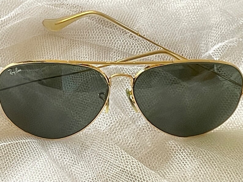 May include: Gold-framed aviator sunglasses with dark green lenses. The sunglasses have a classic teardrop shape and a double bridge. The Ray-Ban logo is visible on the lens. The temples are thin and gold.