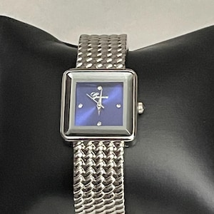 May include: A silver-toned wristwatch with a square blue face and a textured metal band. The watch face has a small diamond accent and a decorative logo. The band is a woven design. The watch is displayed on a black surface.