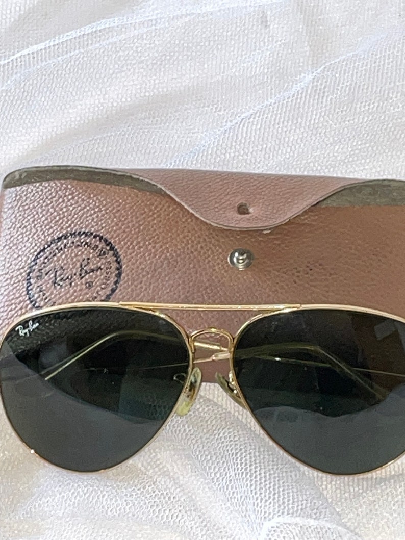 May include: Gold-framed aviator sunglasses with dark lenses and a brown leather case. The case is open, revealing the sunglasses. The Ray-Ban logo is visible on the sunglasses and the case.