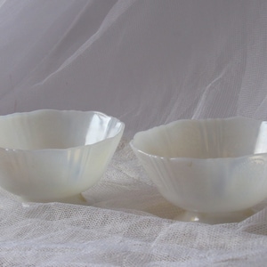 May include: Two opalescent white glass bowls with scalloped rims. The bowls have a pearlescent sheen and sit on small, round bases. The bowls are likely for serving or display.