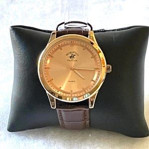 May include: A gold-toned wristwatch with a brown leather band, resting on a black pillow. The watch face features the text "Beverly Hills Polo Club" and "Quartz". The watch has gold hands and a gold bezel.