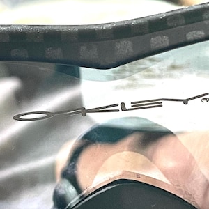 Op de afbeelding: Close-up van een zonnebril met een zwart montuur en donkere lens. Het Oakley-logo is zichtbaar op de lens. Het montuur heeft een textuurpatroon.