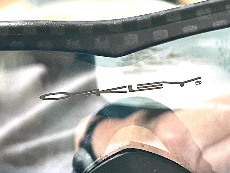 Op de afbeelding: Close-up van een zonnebril met een zwart montuur en donkere lens. Het Oakley-logo is zichtbaar op de lens. Het montuur heeft een textuurpatroon.