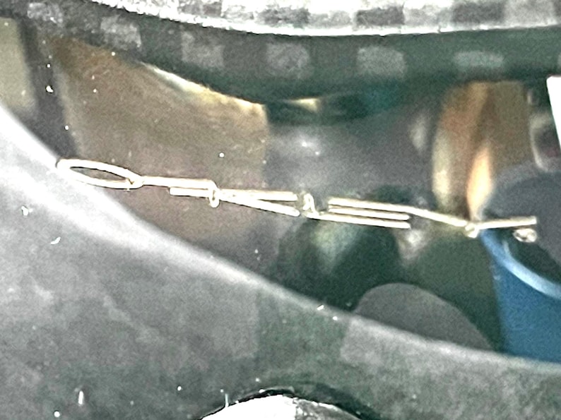 Op de afbeelding: Een zilveren metalen auto-embleem met de letters "D" en "A" zichtbaar. Het embleem is bevestigd op een donkere ondergrond, mogelijk de buitenkant van een auto. De afbeelding is een close-up.