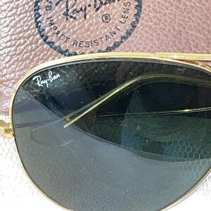 May include: Close-up of Ray-Ban sunglasses with gold frames and dark lenses. The Ray-Ban logo is visible on the lens. The sunglasses are set against a brown leather case with the Ray-Ban logo and text.