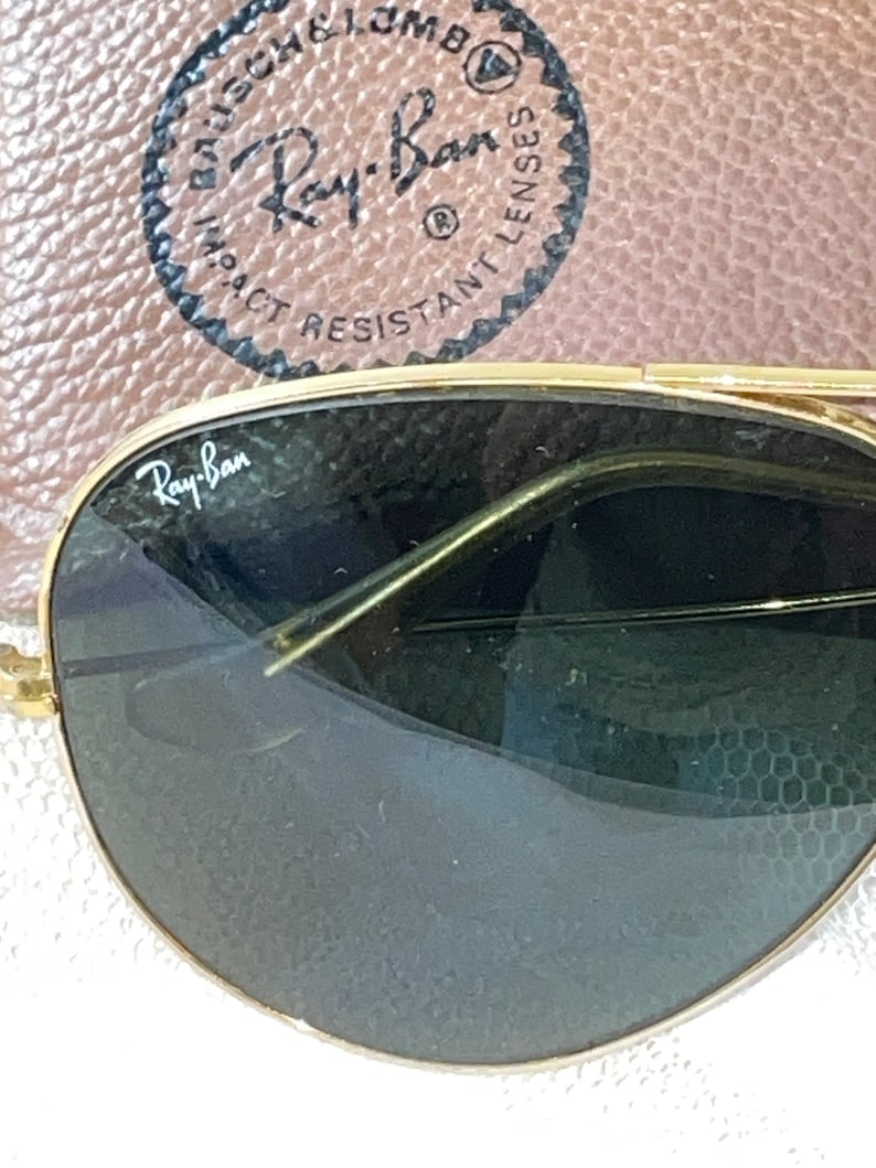 May include: Close-up of Ray-Ban sunglasses with gold frames and dark lenses. The Ray-Ban logo is visible on the lens. The sunglasses are set against a brown leather case with the Ray-Ban logo and text.
