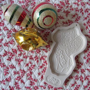 Puede incluir: Un cortador de galletas de &aacute;ngel de cer&aacute;mica blanca con la palabra "Joy" en la parte inferior. El cortador est&aacute; sobre una tela acolchada blanca y roja con tres adornos navide&ntilde;os: uno dorado, uno rojo y verde, y uno rojo y blanco.