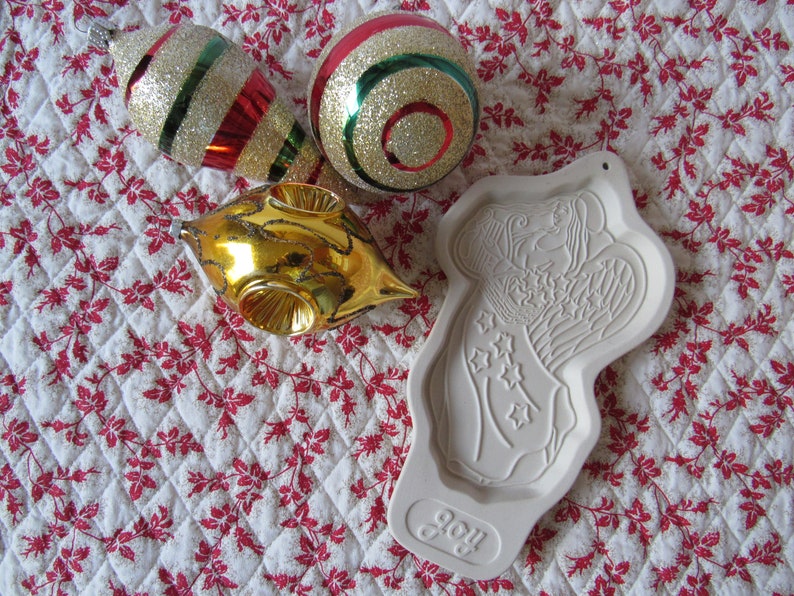 Puede incluir: Un cortador de galletas de &aacute;ngel de cer&aacute;mica blanca con la palabra "Joy" en la parte inferior. El cortador est&aacute; sobre una tela acolchada blanca y roja con tres adornos navide&ntilde;os: uno dorado, uno rojo y verde, y uno rojo y blanco.
