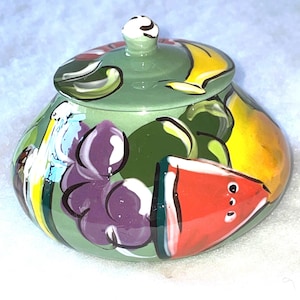 May include: A green ceramic jar with a lid, hand-painted with a colorful fruit design. The jar features a watermelon, grapes, bananas, and other fruits.