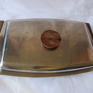 May include: A silver serving tray with a wooden knob on the lid. The tray has a slightly curved shape and a smooth, polished surface.