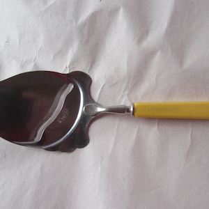 May include: A silver cheese slicer with a yellow plastic handle. The slicer has a wavy blade and a scalloped edge.