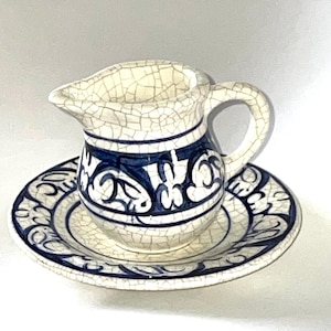 May include: A white ceramic creamer with a blue and white floral design. The creamer has a crackled glaze and a handle. It is sitting on a matching saucer.