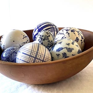 May include: A wooden bowl filled with seven decorative ceramic spheres. The spheres feature blue and white patterns, including floral, plaid, and abstract designs. The bowl is a warm brown color.