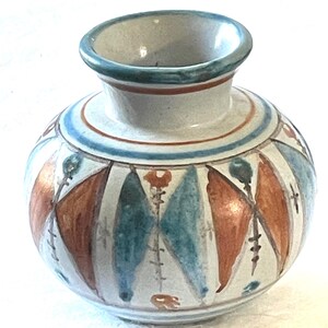 May include: A small, white ceramic vase with a geometric pattern in shades of blue, orange, and brown. The vase has a rounded body and a narrow opening.