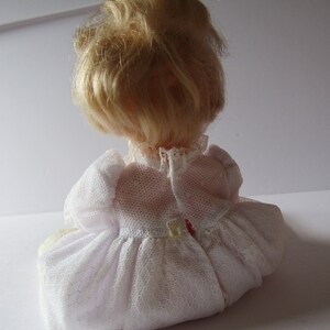 May include: A white porcelain doll with blonde hair wearing a white lace dress. The doll is sitting on a white surface.
