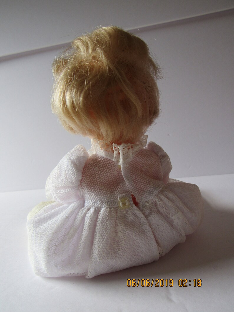 May include: A white porcelain doll with blonde hair wearing a white lace dress. The doll is sitting on a white surface.