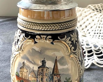 May include: A ceramic beer stein featuring a metallic lid with embossed detailing. The body displays a colorful painted scene of the Ingolstadt Kreuztor city gate, with the text "INGOLSTADT KREUZTOR" printed clearly at the base of the illustration.