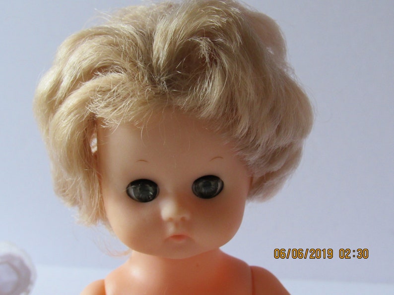 May include: A close-up of a doll's face with blonde hair and large, dark eyes. The doll has a light pink complexion and is wearing no clothing.
