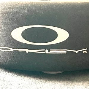 Op de afbeelding: Close-up van een zwarte hoes met het witte Oakley-logo en de merknaam. Het logo is een witte ovaal. De merknaam is in witte, gestileerde letters.