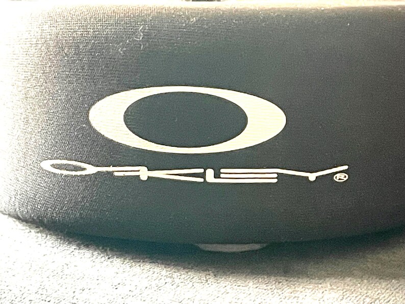 Op de afbeelding: Close-up van een zwarte hoes met het witte Oakley-logo en de merknaam. Het logo is een witte ovaal. De merknaam is in witte, gestileerde letters.