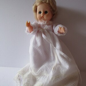 May include: A vintage porcelain doll with blonde hair, blue eyes, and a white lace dress. The doll is wearing a white satin ribbon around its waist.
