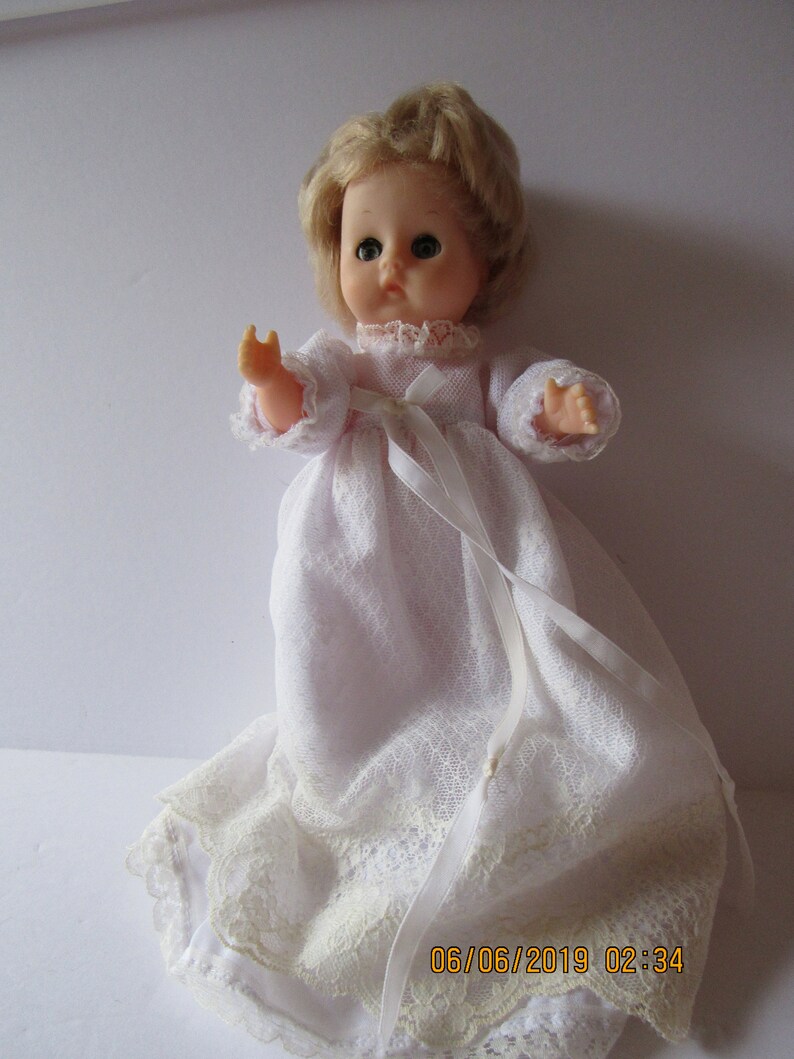 May include: A vintage porcelain doll with blonde hair, blue eyes, and a white lace dress. The doll is wearing a white satin ribbon around its waist.