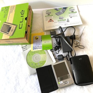 May include: A collection of Sony Clie PEG-TJ25 handheld devices and accessories. Includes a green and brown box, a silver PDA, a black case, a CD, and various cables and manuals. The box has the text "CLIE" and "PEG-TJ25".
