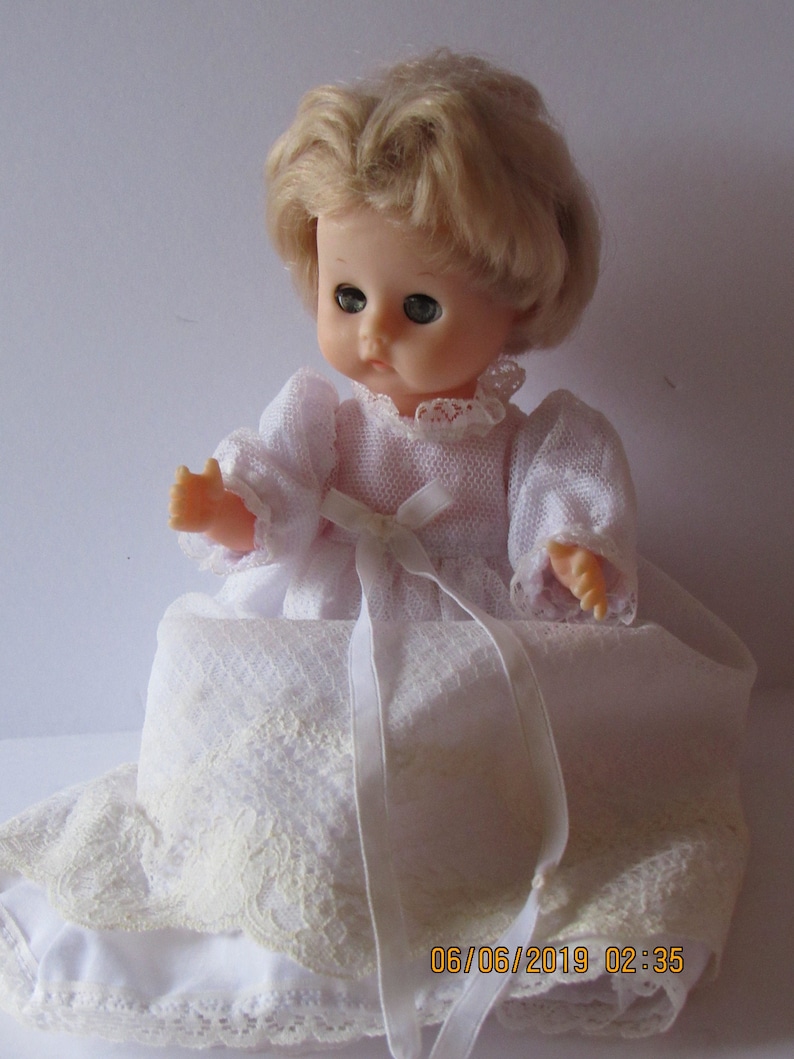May include: A white porcelain doll with blonde hair wearing a white lace dress and a white satin ribbon.