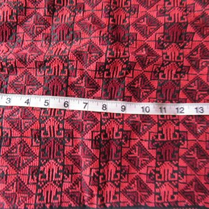 Może przedstawiać: Czerwony materiał z czarnym geometrycznym wzorem. Taśma miernicza umieszczona na materiale, pokazuje pomiar 38 cm.