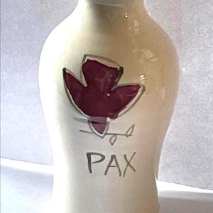 May include: Cream-colored ceramic vase with a burgundy leaf design and the word "PAX" in gray. The vase has a slender neck and a rounded body, suitable for displaying flowers or as a decorative piece.